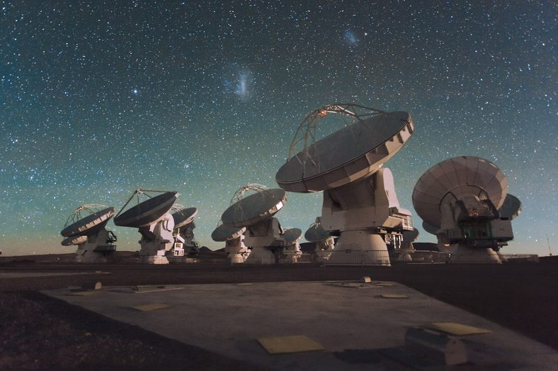 ALMA radio antennas on the Chajnantor Plateau at night, under a sky full of stars, with the Large and Small Magellanic Clouds visible as faint smudges above them.
